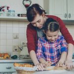 Young attractive mother teaching her little cute daughter rolling dough while cooking together in the kitchen at home on holidays. Family, food and people concept